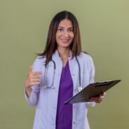 young woman doctor wearing white coat and with stethoscope holding clipboard smiling confident pointing to it with index finger standing over isolated green background